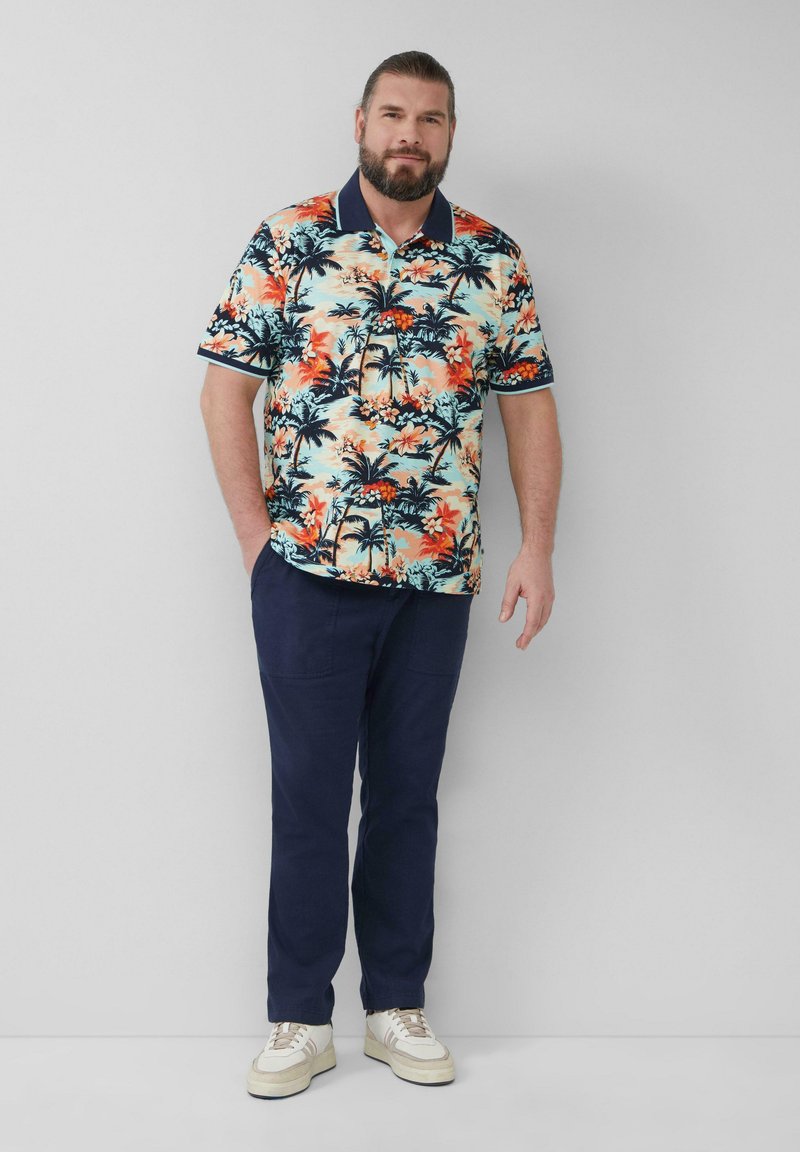 Korte mouwen polo in lichtblauw, met een levendig patroon van bloemen en palmbomen in oranje en zwart. Gepaard met marineblauwe broek en witte sneakers.