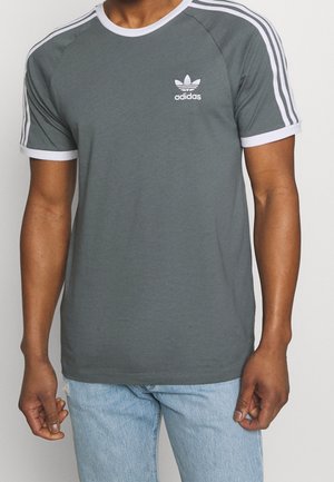 Mann trägt ein graues Adidas T-Shirt mit weißen Abschlüssen und drei weißen Streifen auf den Ärmeln, kombiniert mit hellblauen Jeans.