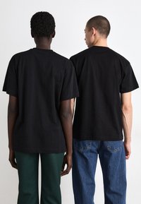 Črne bombažne majice s kratkimi rokavi, pogled s hrbta, nosijo jih dve osebi. Ena jih kombinira z zelenimi hlačami, druga z modrimi kavbojkami.