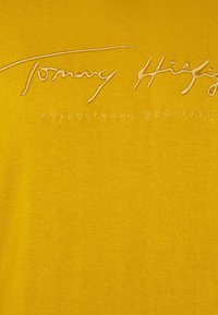 Gelbes Baumwoll-T-Shirt mit geprägtem Text "Tommy Hilfiger" und "ESTABLISHED MCMLXXXV" in einer kontrastierenden Farbe. Glatte Textur.