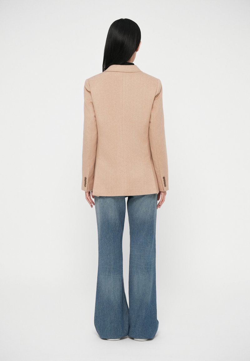 Blazer beige con vestibilità su misura e leggero imbottitura sulle spalle, abbinato a jeans a vita alta svasati, entrambi senza motivi o decorazioni visibili.