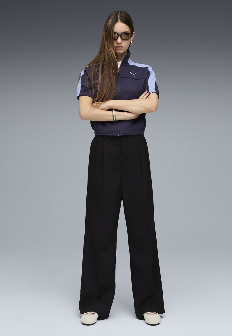 Veste zippée marine avec des accents bleu clair, associée à un pantalon large noir. Portée avec des chaussures blanches et des lunettes de soleil foncées.