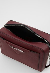 Burgundská taška z texturované kůže se stříbrným zipem, vybavená přední kapsou a vyraženým logem na boku.