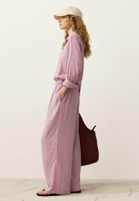Vrouw draagt een roze gestreept playsuit met lange mouwen, een beige pet, zwarte sandalen en draagt een grote donkerbruine handtas, staand opzij.