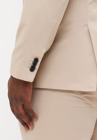 Beige suit jacket sleeve showing textured fabric, four black buttons on cuff, and a close-up of a hand resting on the suit.