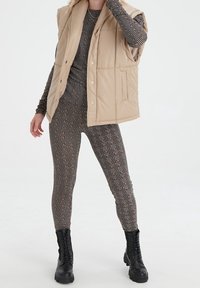 Beige-padded vest with a high collar, paired with fitted leggings featuring a geometric pattern in black and gold. Chunky black boots.