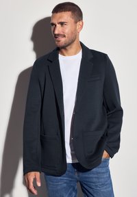 Dunkelgrau Blazer aus weichem Material, mit Reverskragen, zwei Fronttaschen und einer dezenten Brusttasche, kombiniert mit einem weißen Hemd.
