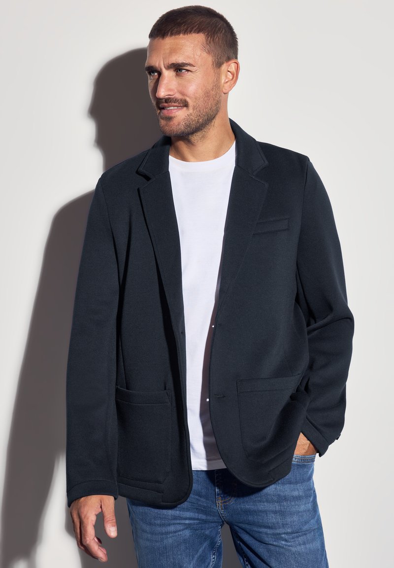Dunkelgrau Blazer aus weichem Material, mit Reverskragen, zwei Fronttaschen und einer dezenten Brusttasche, kombiniert mit einem weißen Hemd.