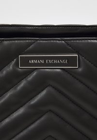 Czarna pikowana torebka ze skóry syntetycznej z srebrną tabliczką z logo "Armani Exchange". Wzór jodełki z widocznymi detalami przeszyć.