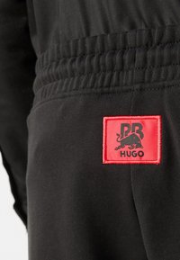 Un pantalon de survêtement noir arbore un patch rectangulaire rouge avec un logo noir et le texte "PR HUGO". La ceinture élastique offre du confort. Tissu lisse et léger.