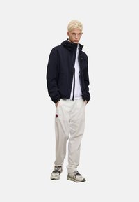 Marine let jakke med hætte, hvid sweatshirt under, beige bukser med logo og grå sneakers med farvede detaljer.