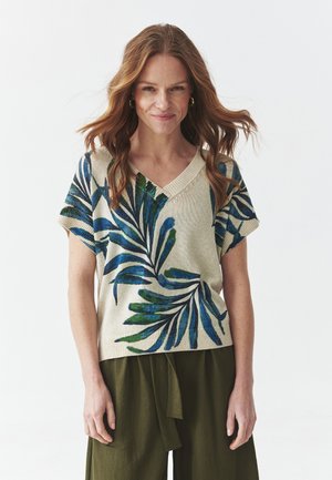 Haut tricot à manches courtes avec un col en V, mettant en avant un motif de feuilles vertes et bleues sur un fond beige clair, en tissu texturé, associé à un pantalon vert.