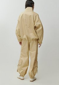 Beige, oversized Jacke mit hohem Kragen und elastischem Bund, kombiniert mit passenden Cargo-Hosen mit seitlichen Taschen und elastischen Bündchen.