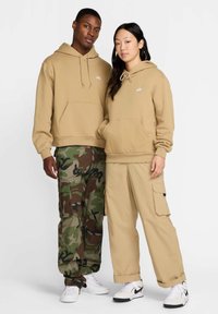 Des sweats à capuche khaki avec poches avant, portés avec un pantalon cargo camouflage. Les deux tenues présentent des coupes amples et un marquage subtil. Tissu texturé visible.