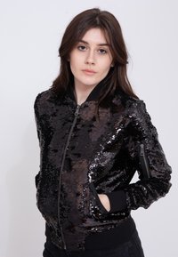 Czarna bomberka z cekinami, z zamkiem z przodu, ściągaczami przy mankietach i na dole oraz kieszeniami bocznymi. Charakteryzuje się błyszczącą, teksturowaną powierzchnią.