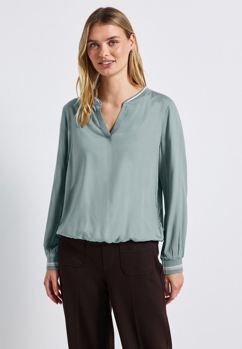 Himmelblaues Blusenshirt mit V-Ausschnitt, langen Ärmeln und elastischen Bündchen. Weicher, glatter Stoff und eine geraffte Taille, kombiniert mit dunklen Hosen.