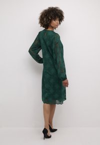 Grön spetsklänning med långa ärmar, blommönster, nyckelhålsdetalj i ryggen och scallop-edged nederkant, parat med svarta högklackade skor.