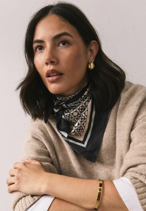 Eșarfă de mătase neagră cu un model geometric în culori discrete, purtată cu un pulover bej. Cercei de aur în formă de cerc și o brățară aurie remarcate.