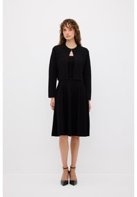 Abito nero al ginocchio con corpetto aderente e gonna svasata, abbinato a un giubbotto nero corto con spalle decorate e scollo a chiave.
