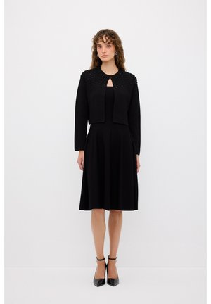 Musta polviin ulottuva mekko, jossa on istuva bodice ja levenevä helma, yhdistettynä lyhythihaiseen mustaan takkiin, jossa on koristeelliset olkapäät ja avonainen kaula-aukko.