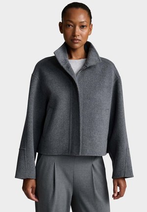 Vrouw draagt een korte grijze wollen jas met hoge kraag en lange mouwen en een bijpassende grijze broek met hoge taille tegen een effen achtergrond.