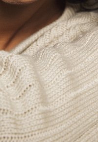 Tissu tricoté crème clair avec motif à torsades en relief, montrant une texture douce et un design drapé le long de l'encolure.