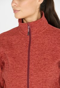 Röd sportjacka med en ståkrage, texturerad tyg och dragkedja. Har ett melerat mönster och kontrasterande dragkedjedetalj.