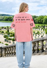 Femme aux longs cheveux blonds portant un T-shirt rose oversize avec texte et graphismes, debout à l'extérieur près d'une balustrade en pierre et d'un jardin.