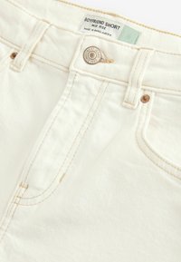 Shorts boyfriend blancs mi-hauteur en denim, avec une fermeture à bouton en métal, des coutures contrastantes, et une étiquette sur la ceinture.