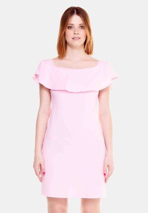 Femme aux cheveux roux mi-longs portant une robe rose clair à épaules dénudées, debout devant un fond blanc uni.