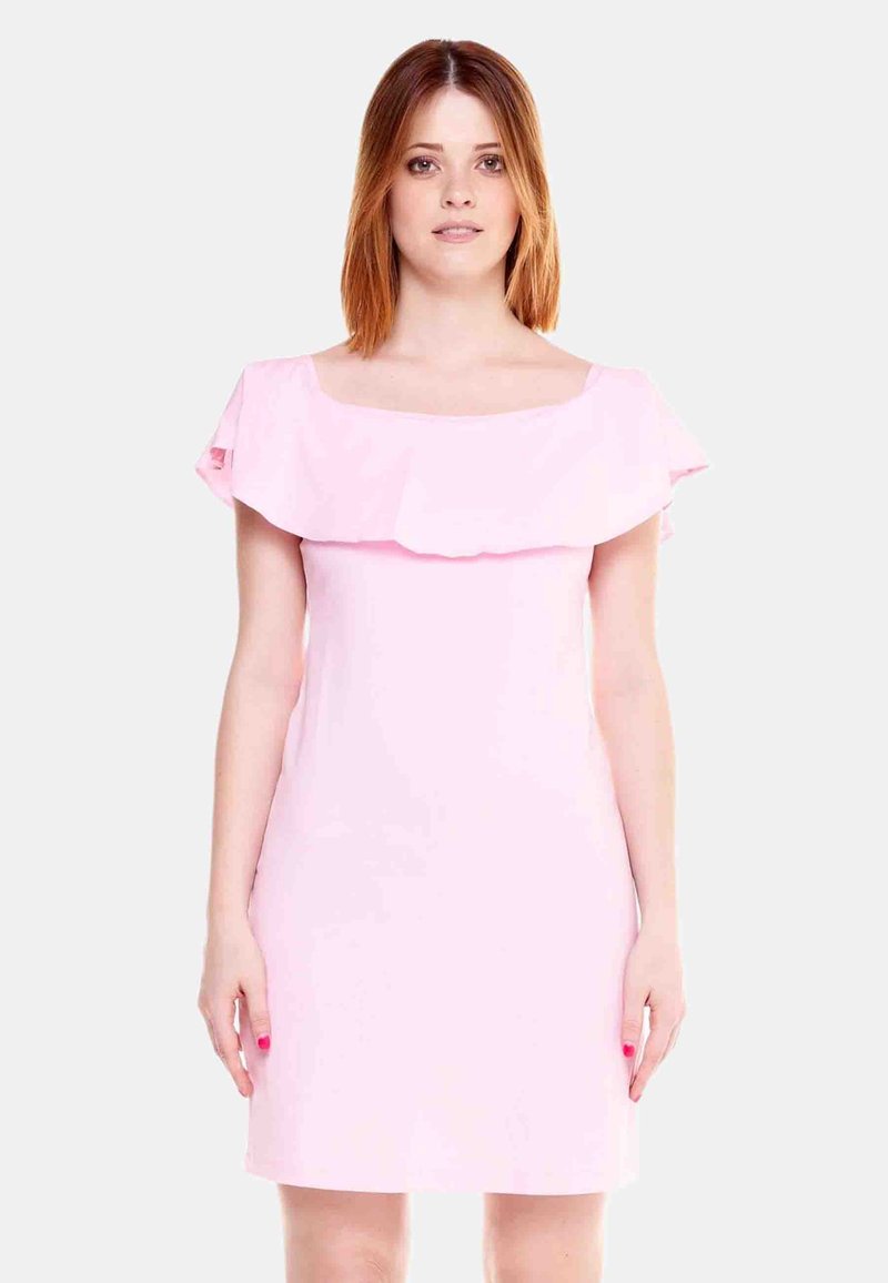 Femme aux cheveux roux mi-longs portant une robe rose clair à épaules dénudées, debout devant un fond blanc uni.