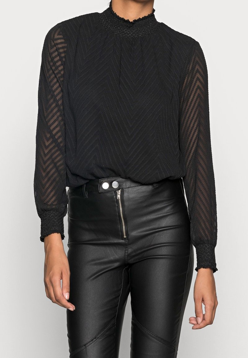 Blouse longue transparente noire à manches longues avec un col haut smocké, présentant un motif chevron subtil et des poignets texturés, associée à un pantalon en cuir noir ajusté.