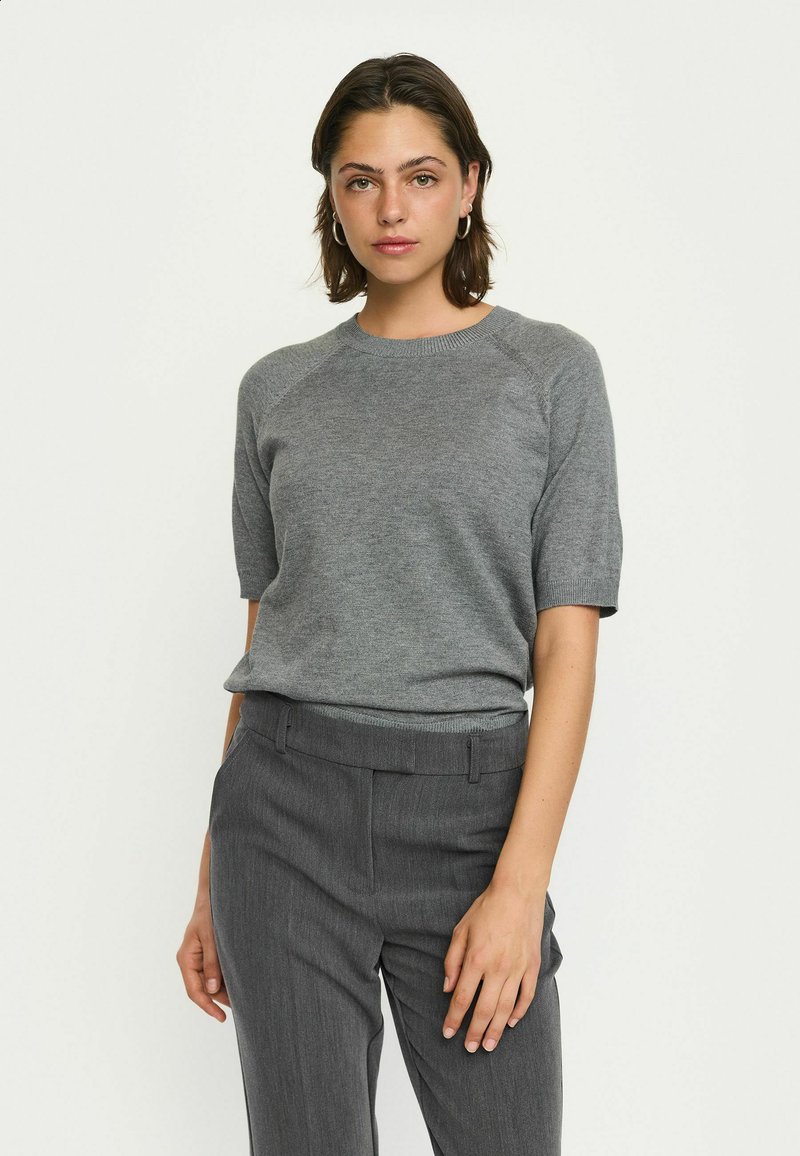 Vrouw die staat in een grijze kortemouwen sweatervest en hoog-waist grijze broek, met haar handen ontspannen naast zich.