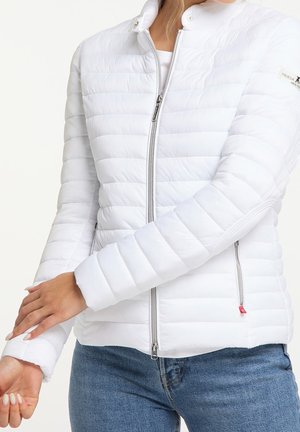 Femme portant une veste matelassée blanche ajustée avec fermeture éclair et un jean bleu, ajustant la manche sur un fond clair uni.