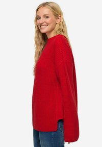 Rode gebreide trui met een textuurafwerking, oversized pasvorm, hoge hals en zijsplitten. Gecombineerd met blauwe denim jeans.
