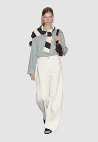 Felpa oversize grigia chiara, jeans bianchi a gamba larga, sciarpa a righe marrone e bianca, tiene una borsa scura, sandali neri. Sfondo semplice.