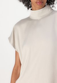 Maglietta leggera color beige chiaro con collo alto, maniche corte, realizzata in morbido tessuto, con una superficie liscia e un design semplice e minimalista.