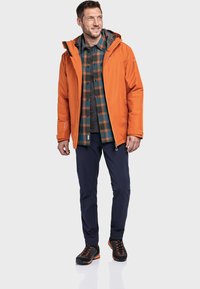 Veste imperméable orange avec une doublure grise, associée à une chemise à carreaux, un pantalon noir et des chaussures noires avec des accents orange.