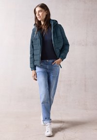 Giubbotto imbottito teal con cappuccio, chiusura con zip e motivo trapuntato; abbinato a jeans azzurri chiari e sneaker bianche. Tessuti lisci in tutto il look.