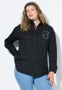 Camicia nera con bottoni e maniche lunghe, dotata di colletto, con una "S" ricamata bianca sul lato sinistro. Indossata con jeans blu.