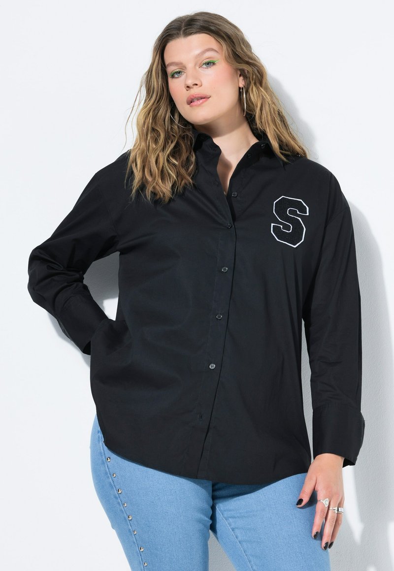 Camicia nera con bottoni e maniche lunghe, dotata di colletto, con una "S" ricamata bianca sul lato sinistro. Indossata con jeans blu.