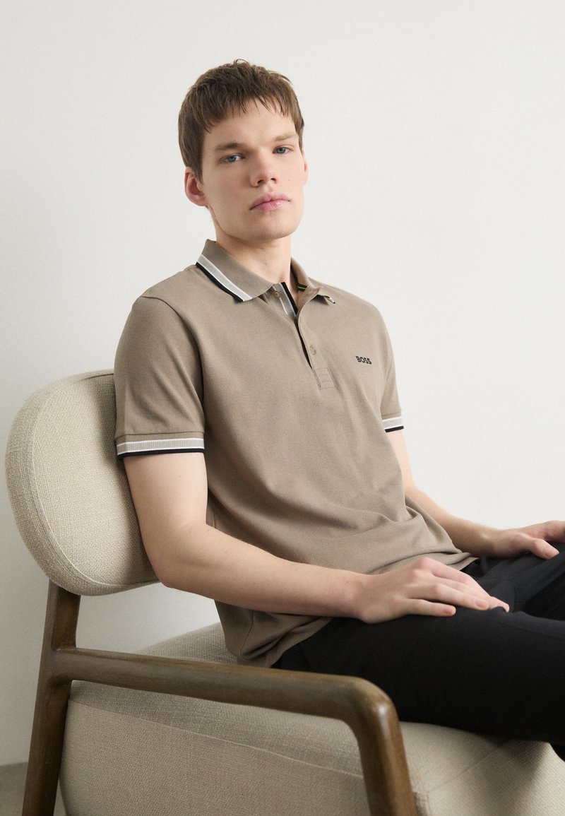 Bež majica s polo ovratnikom, slim fit kroja, s kontrastnim prugastim ovratnikom in izvezenim logotipom. Model sedi na svetli stoli z leseno konstrukcijo.