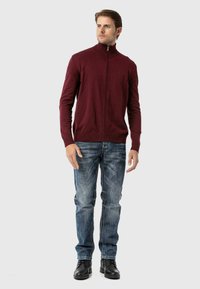 Burgunderfarbener Reißverschluss-Pullover mit geripptem Kragen, kombiniert mit ausgewaschenen blauen Jeans und schwarzen Stiefeln. Schlichtes Design mit einer figurbetonten Silhouette.