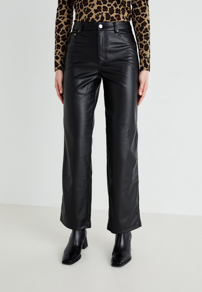 Femme portant un pantalon droit en simili cuir noir, des bottines noires à talons et un haut à manches longues imprimé léopard, debout devant un fond uni.