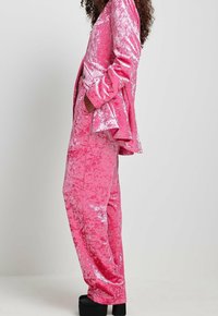 Samtene rosa Anzug mit schwingender Jacke und geraden Hosen. Glänzende Textur, Taschen und maßgeschneiderte Passform. Schwarze Plateauschuhe abgebildet.