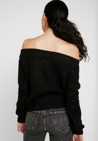 Pull en tricot noir épaules dénudées avec un motif texturé et des poignets côtelés, associé à un jean en denim gris, présentant une coupe décontractée.