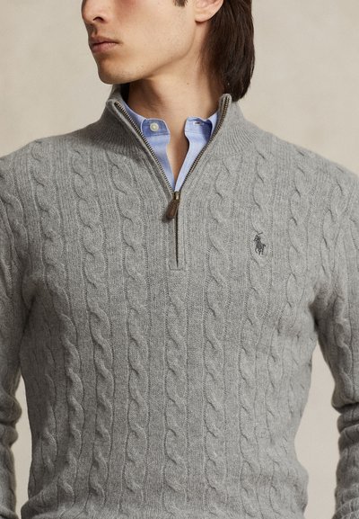 Uomo che indossa una camicia azzurra con colletto sotto un maglione grigio lavorato a trecce con mezza cerniera e un piccolo logo ricamato sul petto.