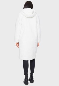 Manteau long blanc et duveteux avec capuche, présentant une surface texturée et des manches longues, assorti à des leggings noirs et des bottines.