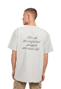 Hvid kortermet t-shirt med en løs pasform, der har sort kursiv tekst på ryggen, som siger: "Det er okay at unfollow folk i det virkelige liv."