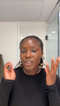 Młoda kobieta z warkoczem, ubrana w czarną koszulę z długim rękawem i duże kołowe kolczyki, wykonująca gesty rękami w dobrze oświetlonej łazience.